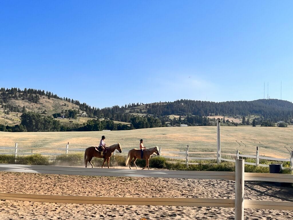 Two riders on horseback in the outdoor riding arena at Kiesling Acres in Valleyford, Washington, with rolling hills in the background.
